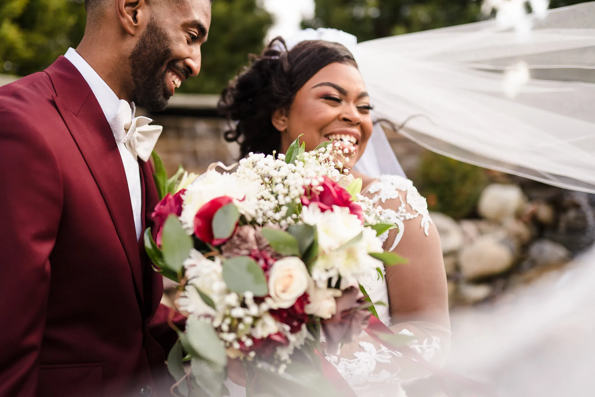 Bride laughing with her bouquet beside her groom in a burgundy suit