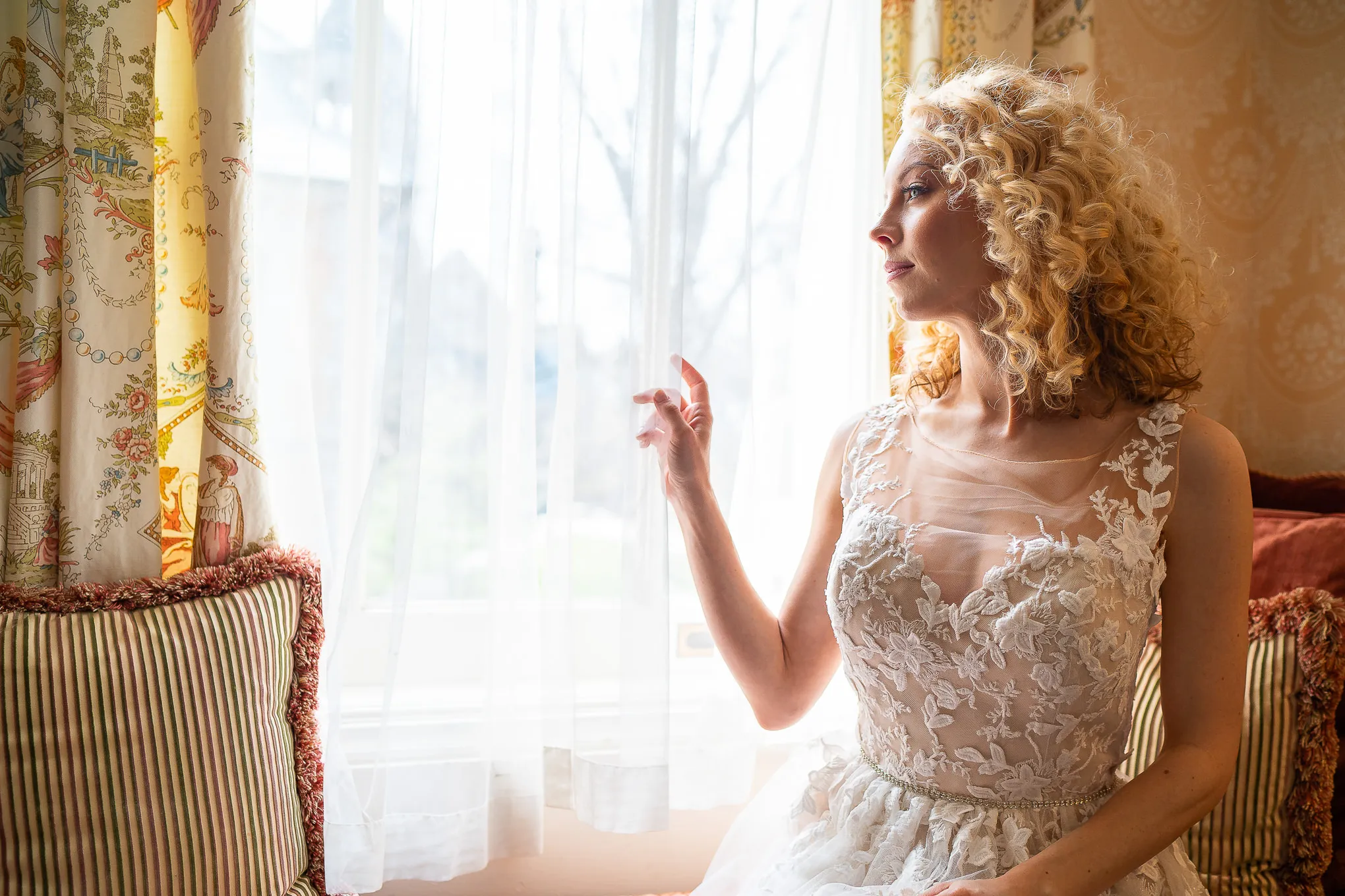 Bride in a lace gown sitting by a sunlit window