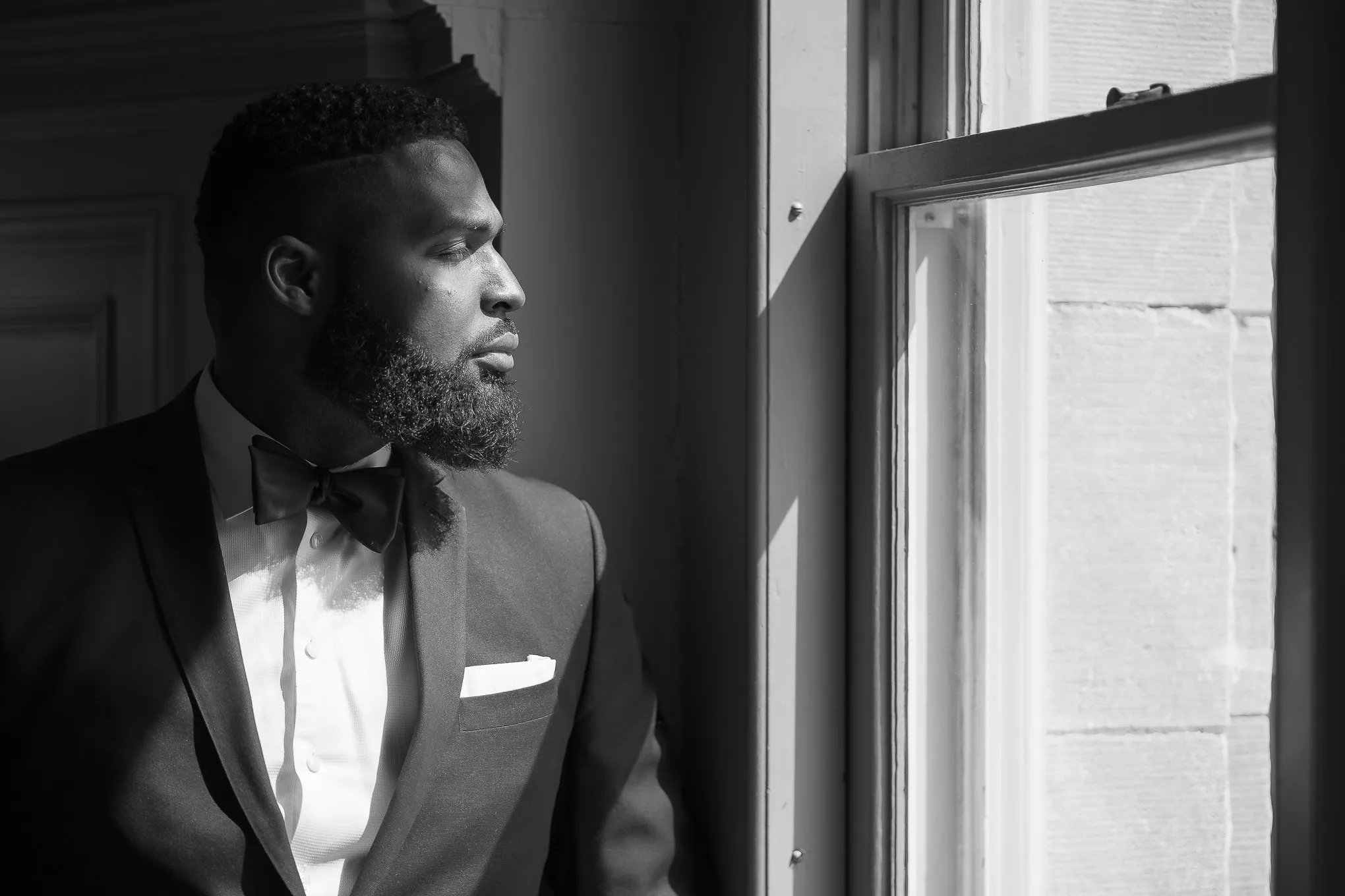Black and white portrait of a groom in a tuxedo looking out a window