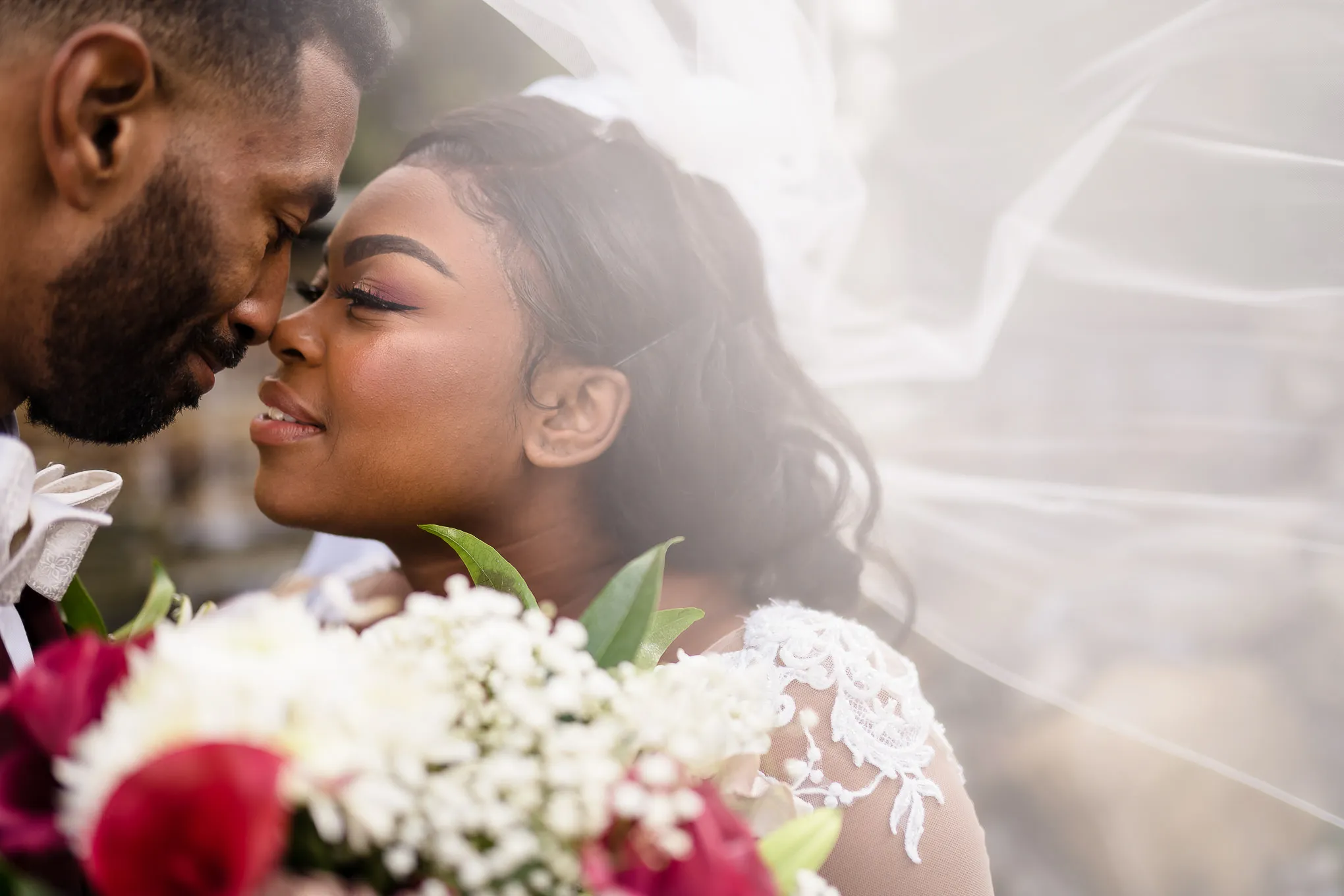 Couple nose to nose under her veil, holding a red and white bouquet